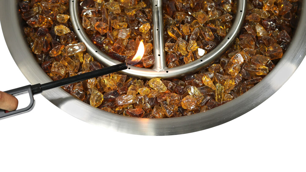 A close-up view of a grill lighter holding a flame to a match-lit stainless steel burner with amber-colored fire gems.